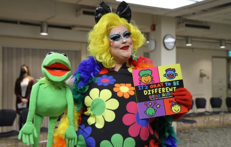 A drag queen in a yellow wig and brightly coloured clothing holds a frog puppet and a book.