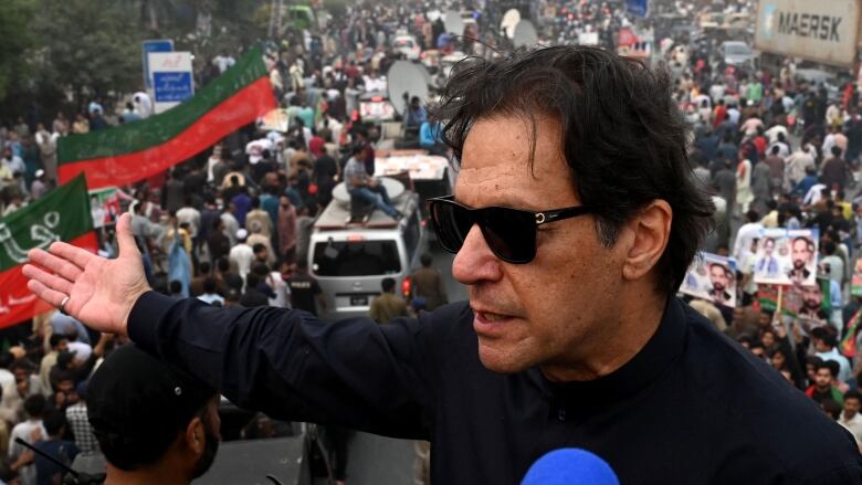 A man with dark hair and sunglasses stands on top of a vehicle and gestures to a large crowd in the street behind him. 