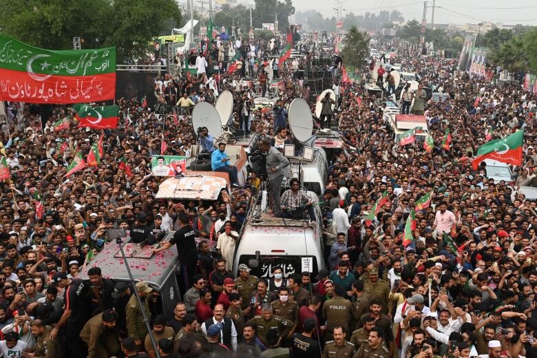 Huge crowds of people waving flags surround vehicles during a political rally. 