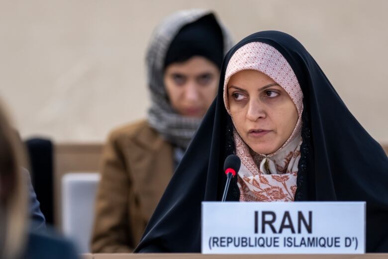 A woman wearing a head covering speaks from a desk, with a title card in front of her reading 'Iran.'