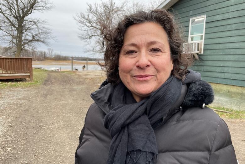 A woman, Linda McVicar, stands outside in front of the lake with a green cabin behind her and smiles at the camera.