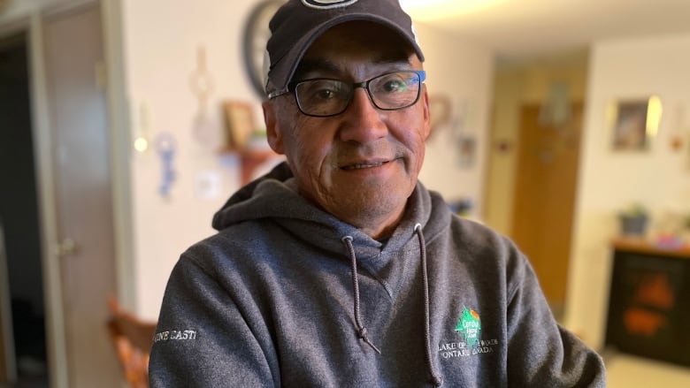 A man wearing a hat, glasses and a grey sweater crosses his arms and looks at the camera.