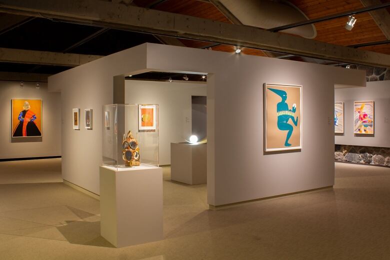 Photo of a gallery at the McMichael Canadian Art Collection. The room is dimly lit and a selection of colourful figurative paintings hang on grey walls. An ornate gas mask is encased in plexiglas on a plinth at the centre of the room. Behind it is a three-walled gallery within the gallery containing an orb-shaped lamp, part of a sculptural work of art.