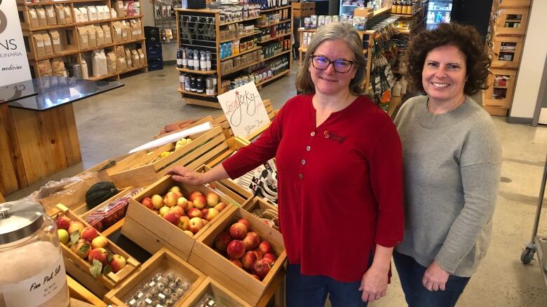 Two women standing in a small store near some produce.