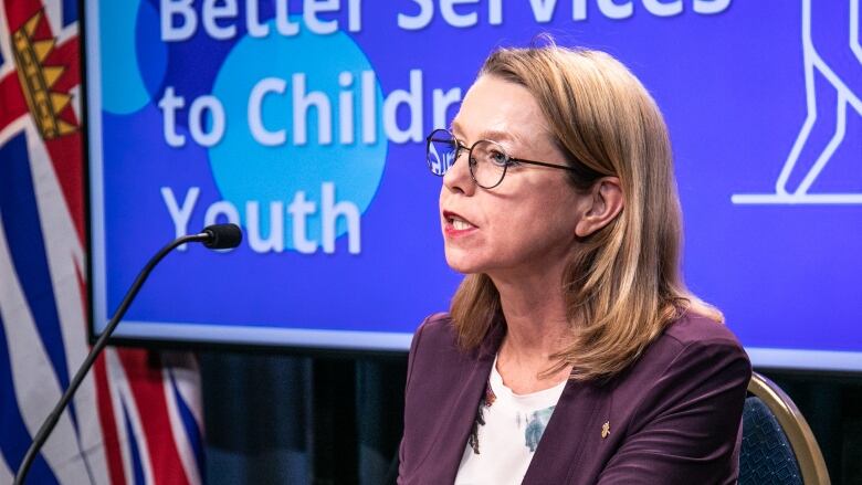 A white woman wearing a purple coat addresses a news conference, with a slide behind her reading 'Better services to children and youth.'