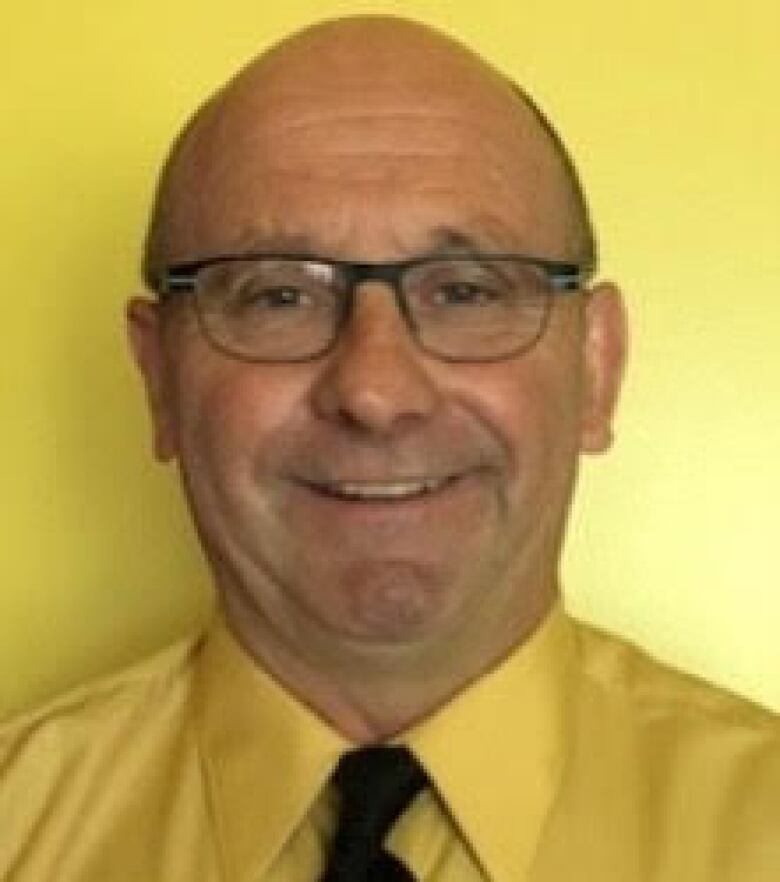 A bald man with glasses wears a yellow shirt and a tie and smiles.