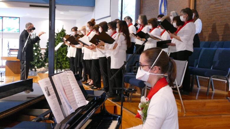 Person playing piano in foreground, choir and conductor behind, all wearing masks.