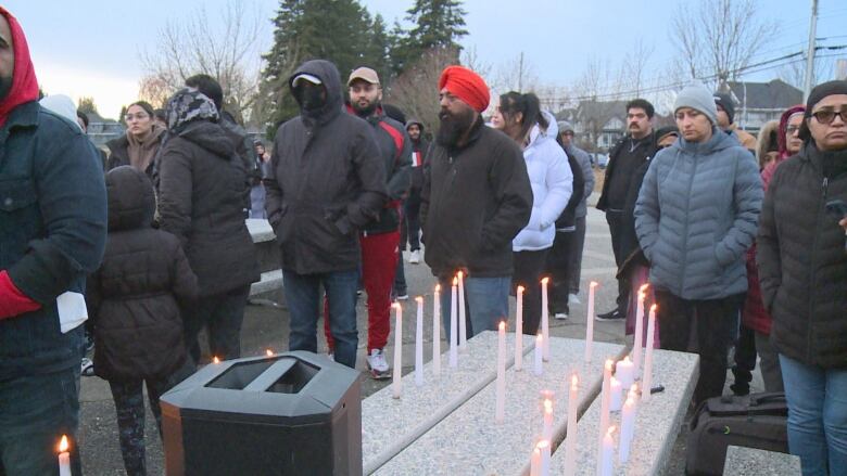 People gather around a bench that is filled with candles at an outdoor vigil.