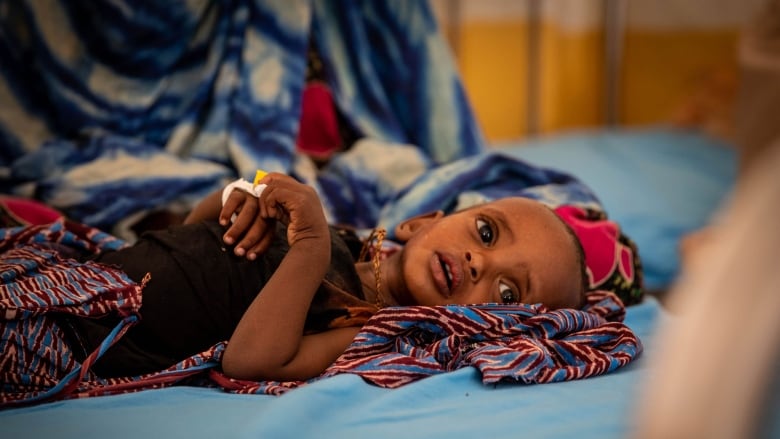 A wide-eyed child lies on blue sheets with some colourful patterned fabrics around her.