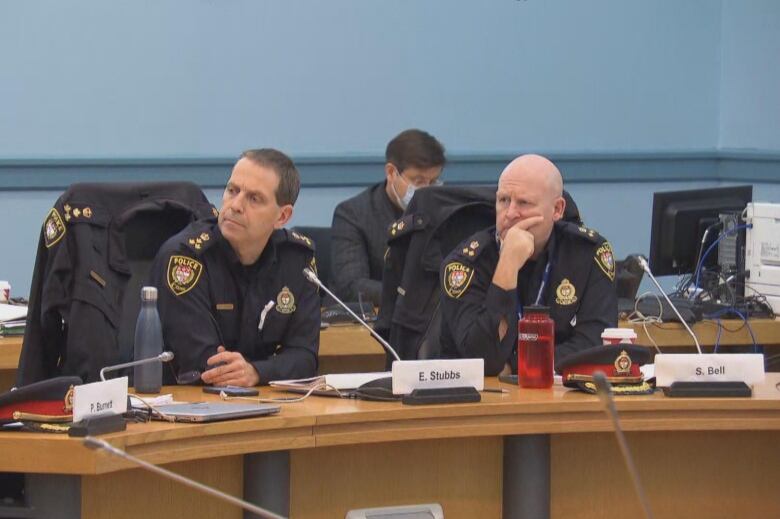 Two police officers look to their right and listen from a desk in a meeting room.