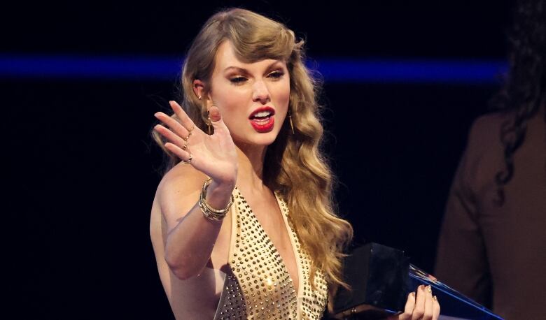 Taylor Swift, a blonde woman wearing a gold dress, waves at an awards ceremony.