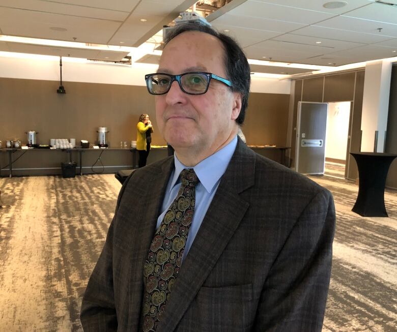 A person in a dark plaid suit jacket wearing glasses stands in a room with a neutral colour palette.