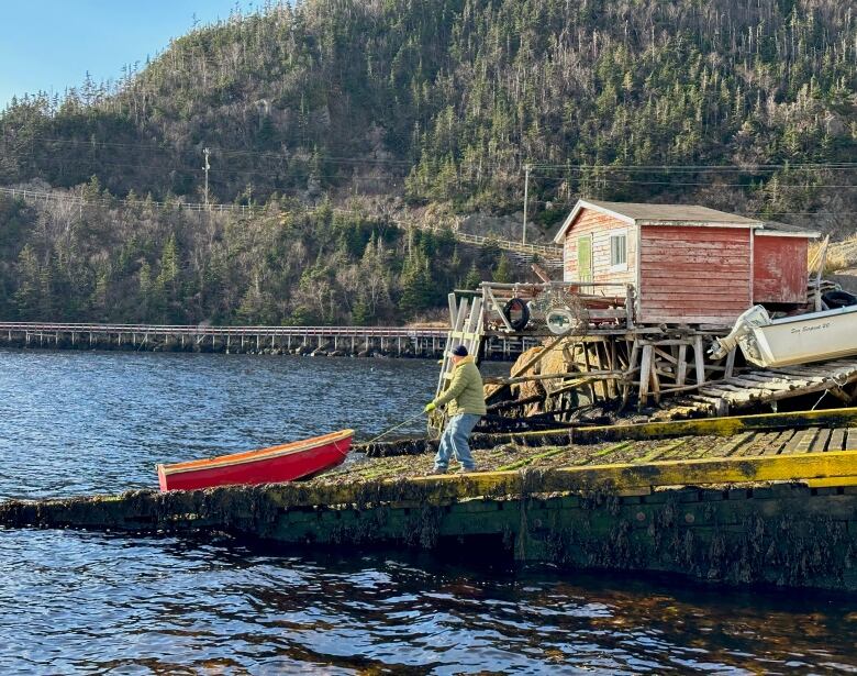 A wide shot of a man pulling a small boat up a slipway, contrasted with a scenic harbour