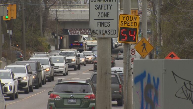 Cars are seen along a busy stretch of road, with signs in the foreground. 