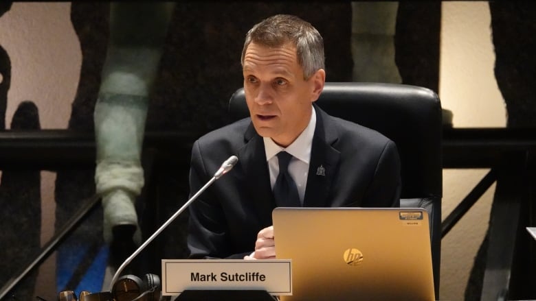 A man in a suit sits in front of a microphone and a laptop and delivers a speech.