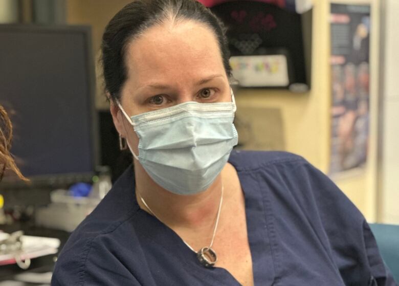 A nurse sits looking at the camera. She is wearing scrubs and a medical mask