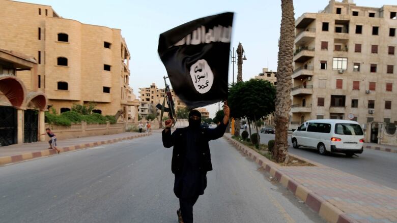 A person dressed all in black with much of their face covered holds a rifle in one hand and a black-and-white Islamic State flag in the other, walking alone in the middle of a street in Raqqa, Syria.