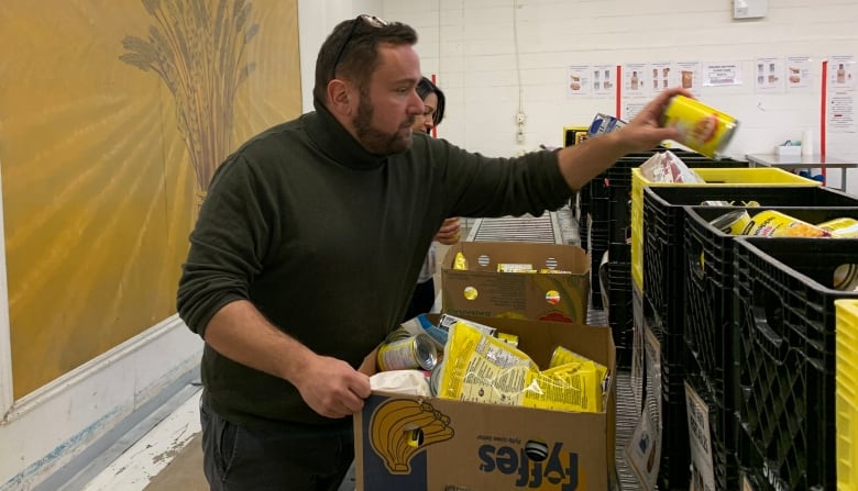 A man in a warehouse stands in front of a row of bins and boxes. He reaches toward one with a can of food in his hand.