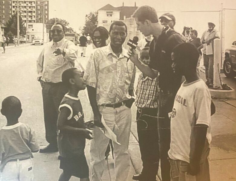 A reporter holds a microphone while interviewing a man on a street as he is surrounded by four boys and a woman.