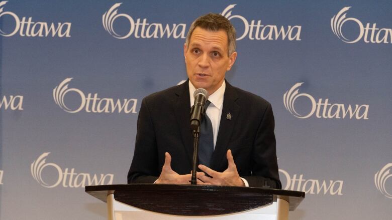 A man wearing a suit speaks at a podium in front of a blue background with the word Ottawa written on it.