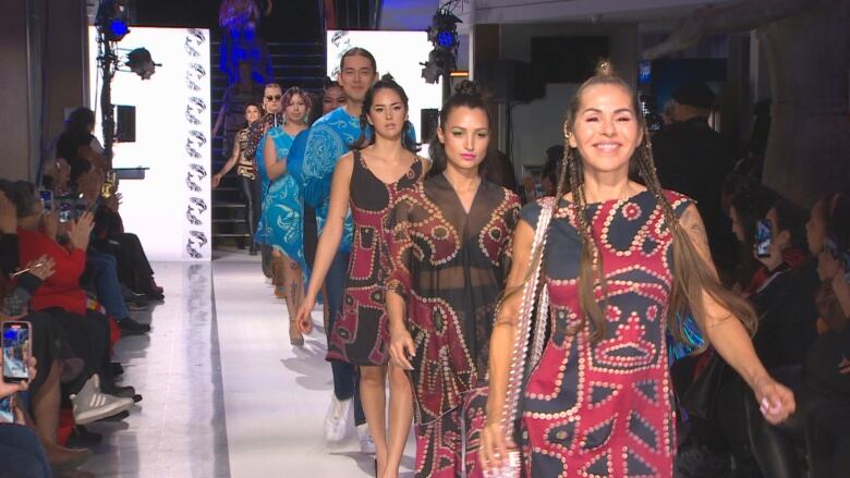 Models walk down runway wearing red and blue, smiling at the end of a collection showing.