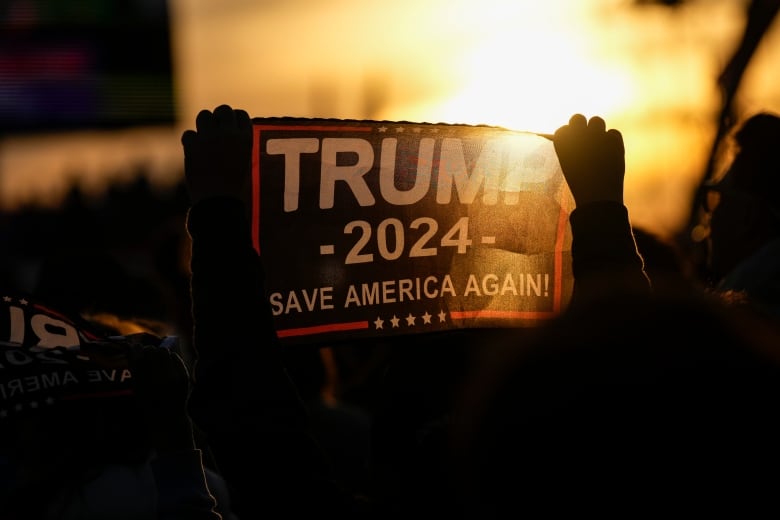 A shot at sunset of a crowd waving banners that say Trump 2024, Save America Again.