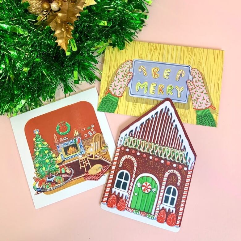 Flat-lay photo of three illustrated greeting cards, shot on a pink surface. The cards depict Xmas scenes: a baking tray with cookies that spell 