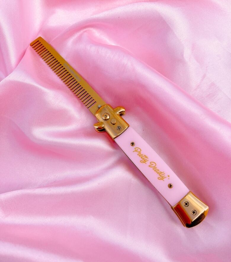 Photo of a pink and brass switchblade comb, shot on a backdrop of bubblegum pink satin. The handle reads in gold script: 