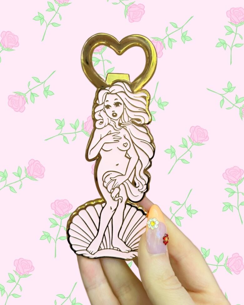 A hand with a flower manicure holds a bottle opener shaped like the goddess Aphrodite standing on a shell. It is in pale pink enamel and the bottle opener appears to be brass. The background is a digital illustration of repeating pink roses.