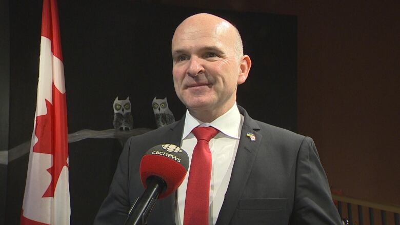 A bald man in a dark suit and a red tie stands in front of a microphone.