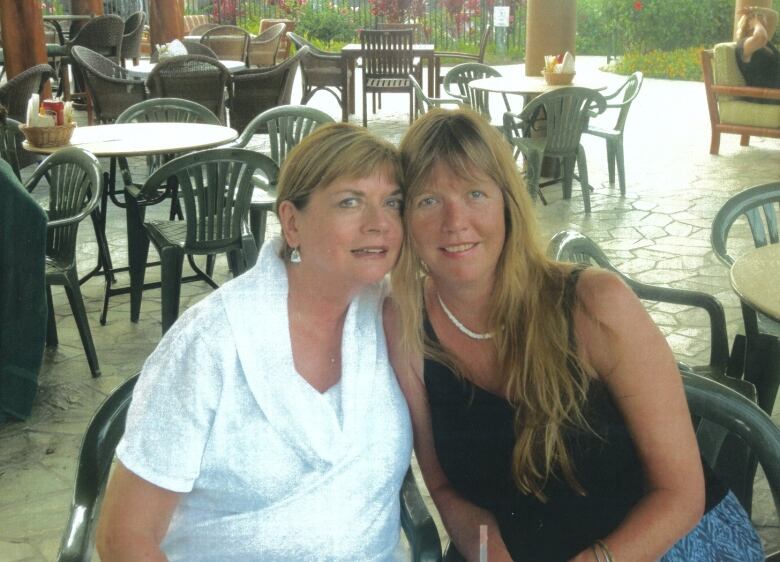 Two women sit on a patio together with tropical plants in the background.