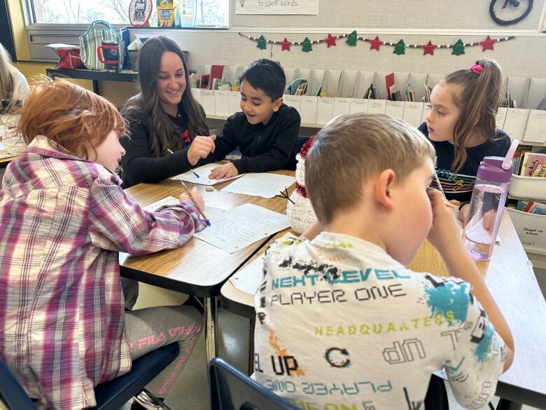 A young teacher with brown hair sits at a table with four students. 