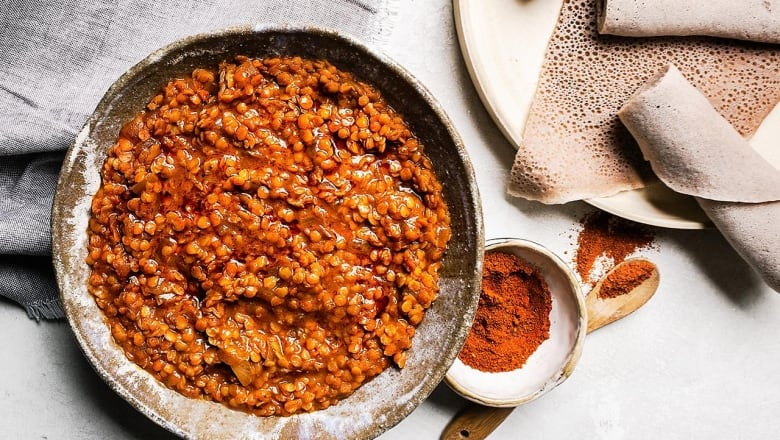 overhead shot of a bowl of lentil stew with a small bowl of spices sitting next to it. a plate with flatbread (injera) sits next to that. 