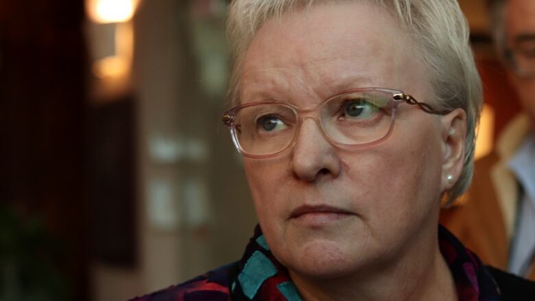 A woman with short grey hair and glasses glances to the left of the photo with a serious expression. It's fairly close-up and only the collar of a black blouse with aqua squares and some red patterning is visible.