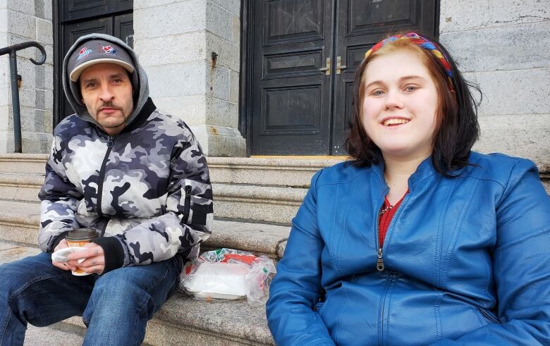 A man wearing a baseball cap, a camo sweater and jeans sits next to a woman wearing a bright blue coat and a multi-coloured headband. They're both sat on the steps of the Basilica Cathedral in St. John's. A takeout container is placed between them.