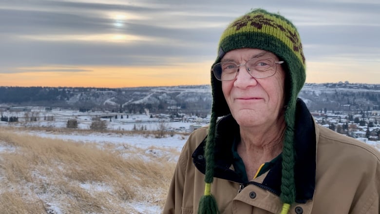 A man wearing a tuque smiles at the camera while behind him you can see snow on the trees in the river valley.