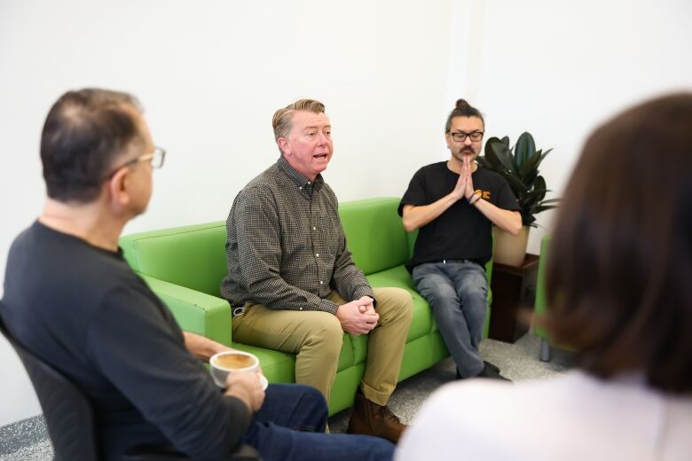 A man sits on a couch surrounded by other people who are listening.