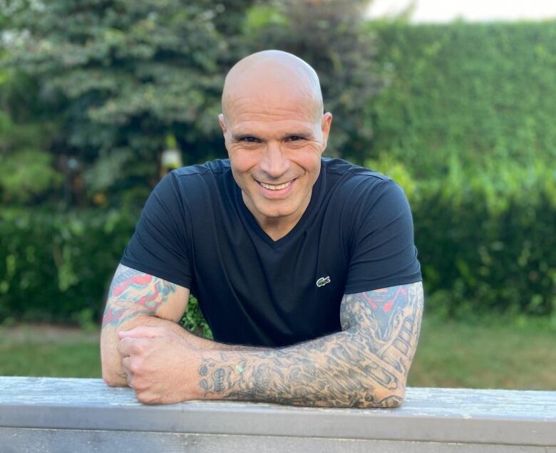 A bald man with tattoos smiles in an outdoor environment.