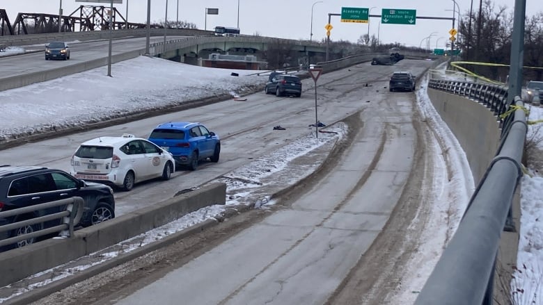 Cars are lined up on a road leading up to a bridge, where one car can be seen smashed into a guardrail. Rurther down the bridge, the wreck of another car can be seen.