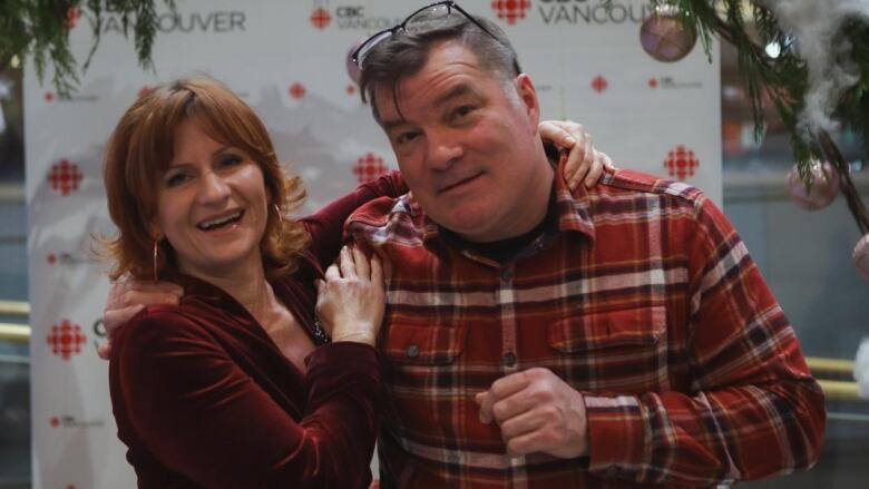 Amy Bell and Stephen Quinn stand with their arms around each other's shoulders. She has red hair and wears a red velvet dress. Stephen wears a red plaid shirt and a smile.