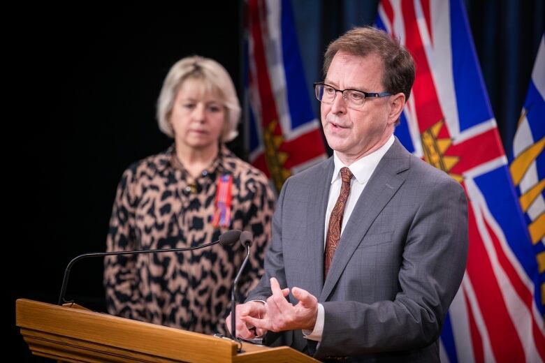 A man speaks at a podium, with the flag of British Columbia behind him. A woman with short hair stands in the background.