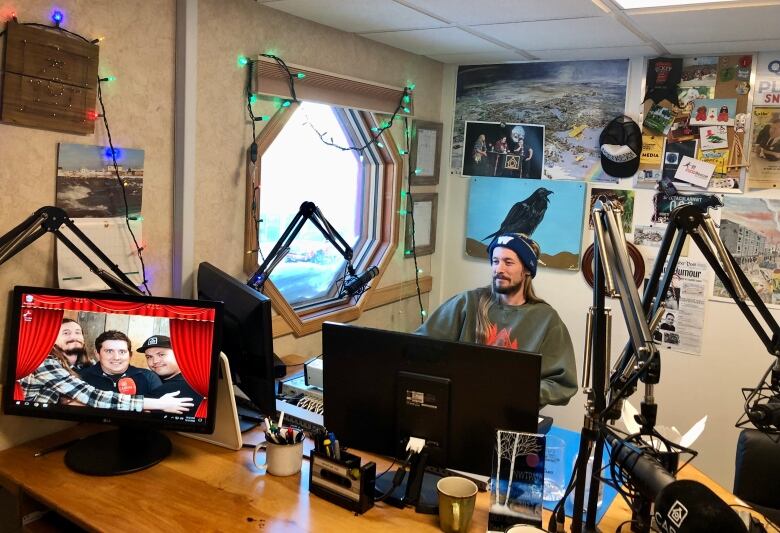 A man in a toque sits at a microphone inside a radio studio, surrounded by computer monitors.