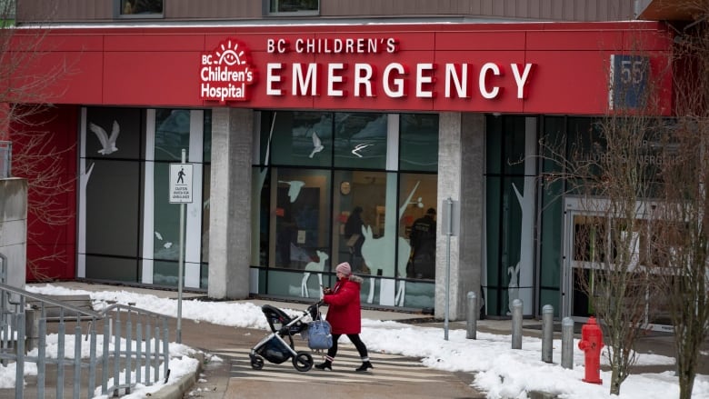 A person in winter clothes pushes a stroller across a pedestrian walkway in front of a building with a sign that reads 'B.C. Children's Emergency.'