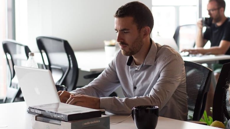 A white man with trimmed facial hair and a collared shirt types on a laptop.