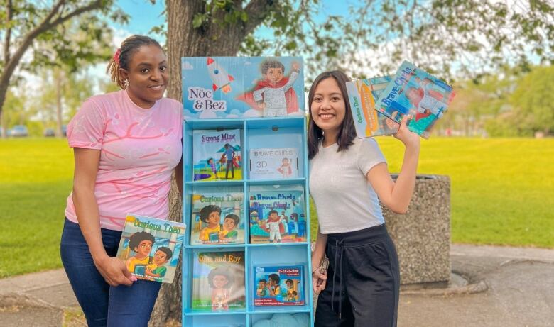 Two smiling women holding books in a park. They stand on opposite sides of a display case containing more children's books. 