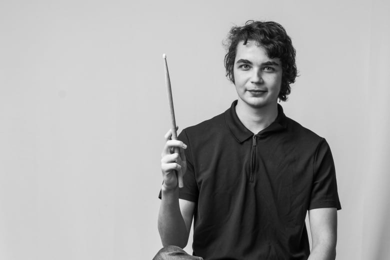 A young man smiles slightly and holds a drumstick vertically, with his elbow resting on his knee.