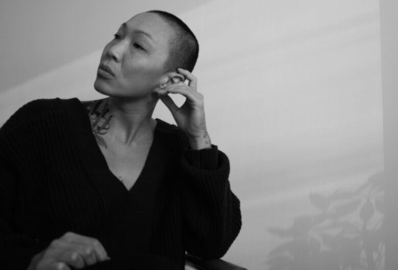 An artistic black-and-white photo shows a portrait of a woman with close-cropped hair, sitting with her head resting on her hand and her head tilted to the right. A beam of sunlight casts the shadow of a plant on the wall behind her.