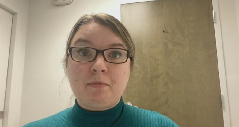 A woman wearing glasses and a teal shirt looks into the camera.