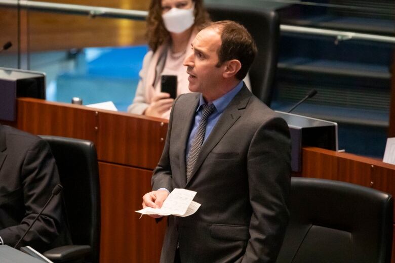 A man in a dark suit speaks on the floor of city council.