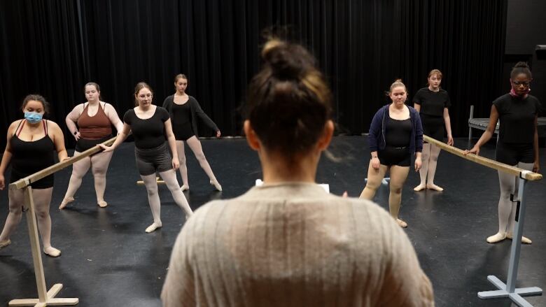 A woman with her hair in a bun stands in front of a group of seven dancers. Four are on one side and three are on the other. They're all wearing ballet attire and ballet shoes. 